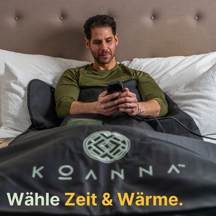 Braunhaariger Mann mittleren Alters liegt mit der schwarzen Saunadecke und einem Buch im Bett und bedient gerade den Controller.